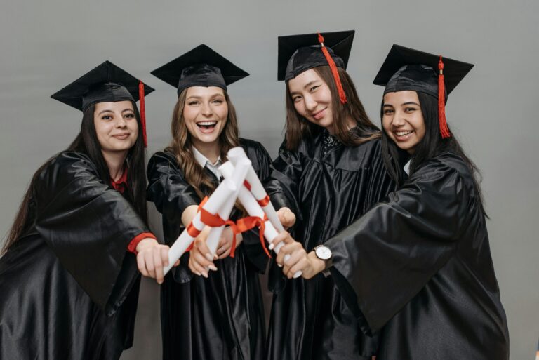 A joyful group of multiracial women in graduation gowns holding diplomas.